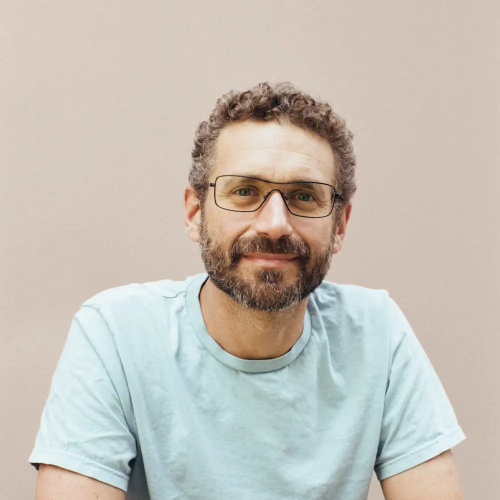 A photo of Ilan Gur smiling and sitting down in front a cream background.