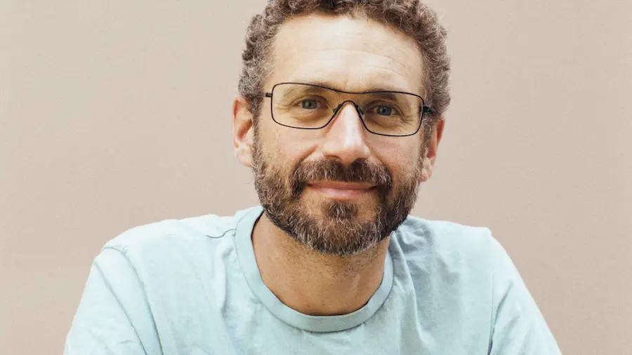 A photo of Ilan Gur smiling and sitting down in front a cream background.