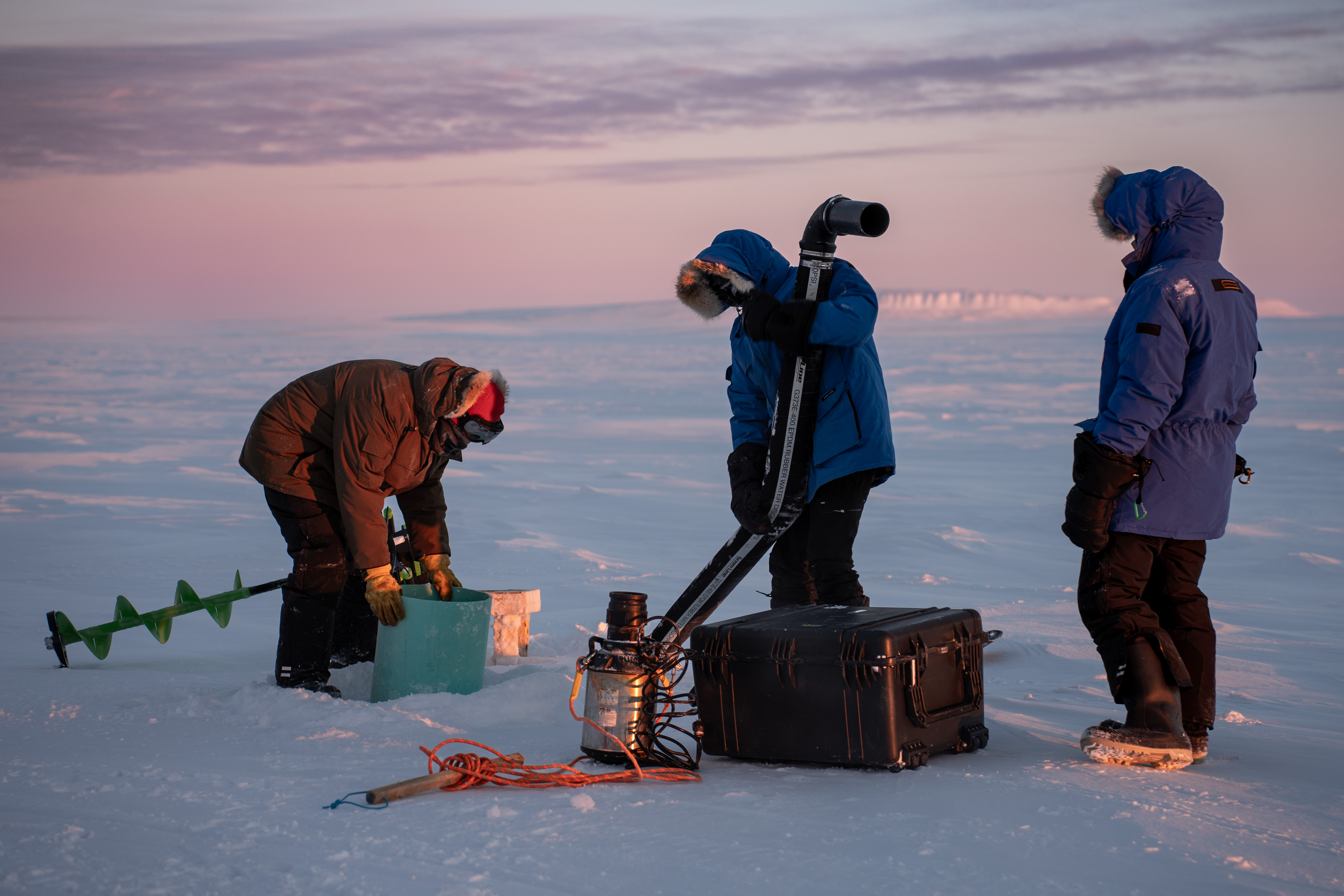A team of three people dressed in warm winter clothes working on a water pump on ice fields in Canada