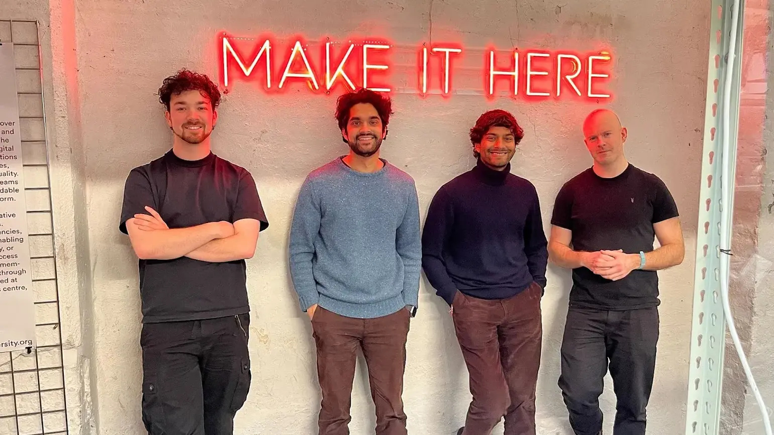 Four men standing in front a wall with a neon sign on the wall behind them saying 'Make it here'