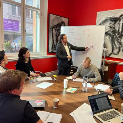 A photo of a group of people sitting around the table and looking at davidad writing on a whiteboard.