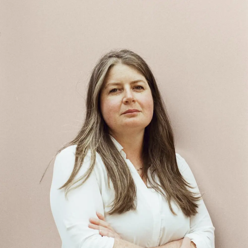 A photo of Tammy Thomas Brown with her arms crossed standing against a cream background. 