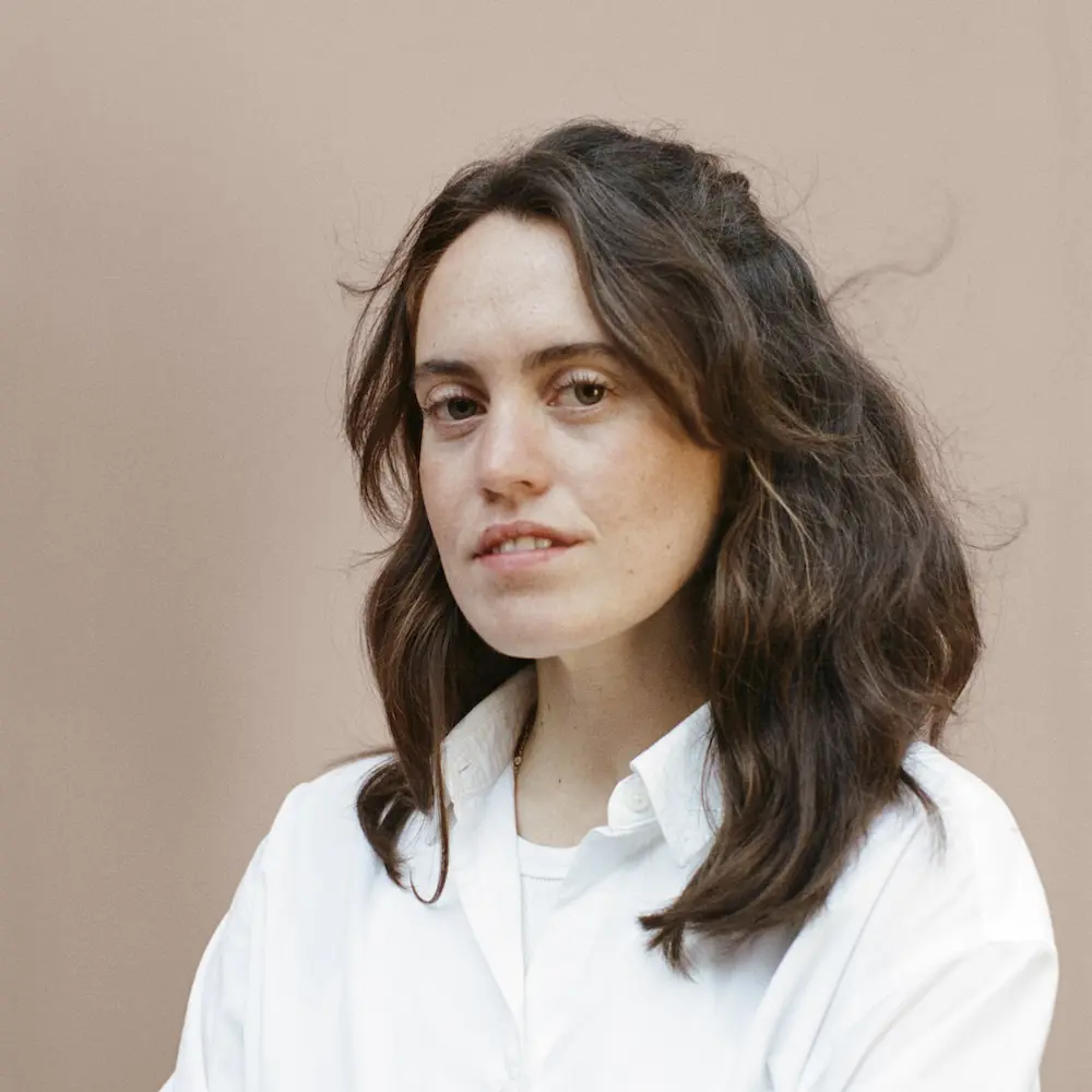 A photo of Pippy James with her arms crossed standing against a cream background. 