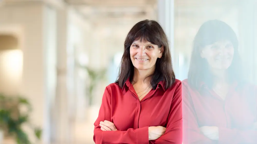 A photo of a woman, Kathleen Fisher, leaning against a glass wall and smiling at the camera with her arms folded.