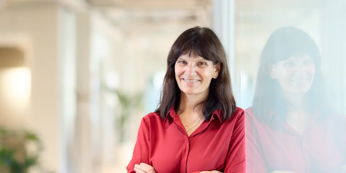 A photo of a woman, Kathleen Fisher, leaning against a glass wall and smiling at the camera with her arms folded.